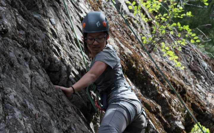 an outward bound student wearing safety gear looks down as they scale a rock wall